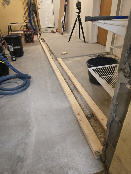 Förrådsgolv med träreglar längs en ränna, laser på stativ, olika arbetsredskap i bakgrunden. Renovation pågår.