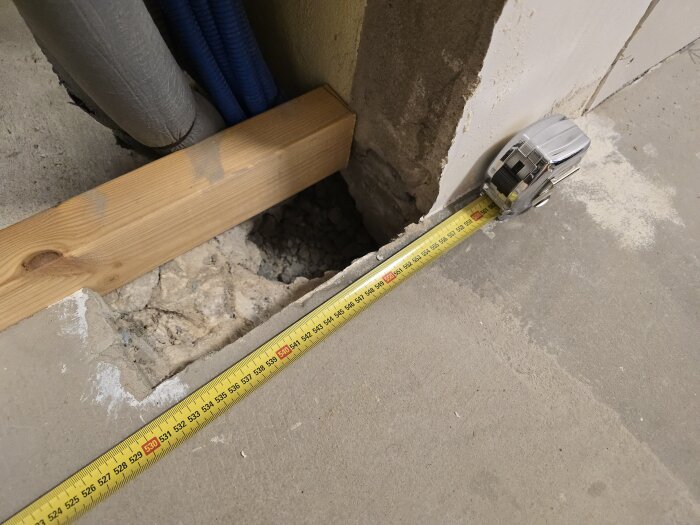 Måttband mäter avstånd vid en vägg med träregel och ventilationsrör synliga, bredvid en skåra i betonggolvet under renovering.