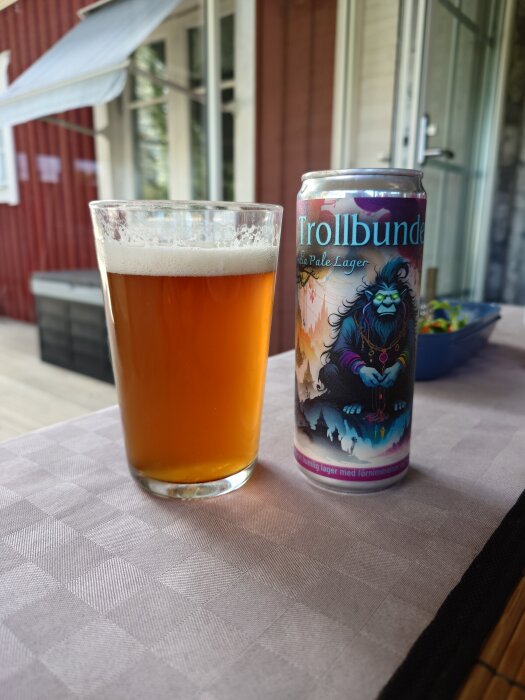 Ett glas med grumlig öl och en burk "Trollbunden Pale Lager" står på ett dukat bord utomhus vid en röd träbyggnad.