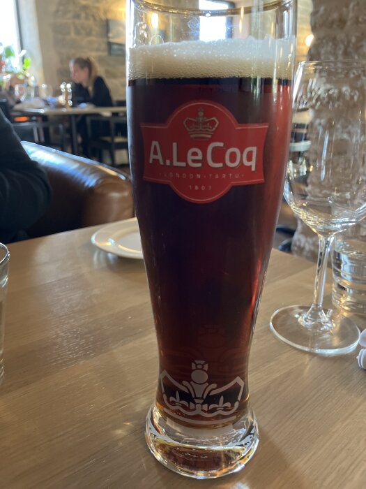 Hög glas fylld med mörk öl, A. Le Coq-logga synlig, placerad på ett träbord i en restaurangmiljö med suddig bakgrund.