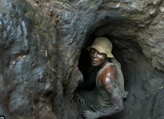 En person i smutsiga kläder och hatt arbetar i en trång gruvgång, omgiven av jord och sten.