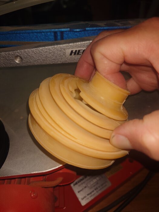 Hand håller en 3D-utskriven del av TPU, formad som en bälg, mot en maskin i bakgrunden, eventuellt till en äldre häcksax.