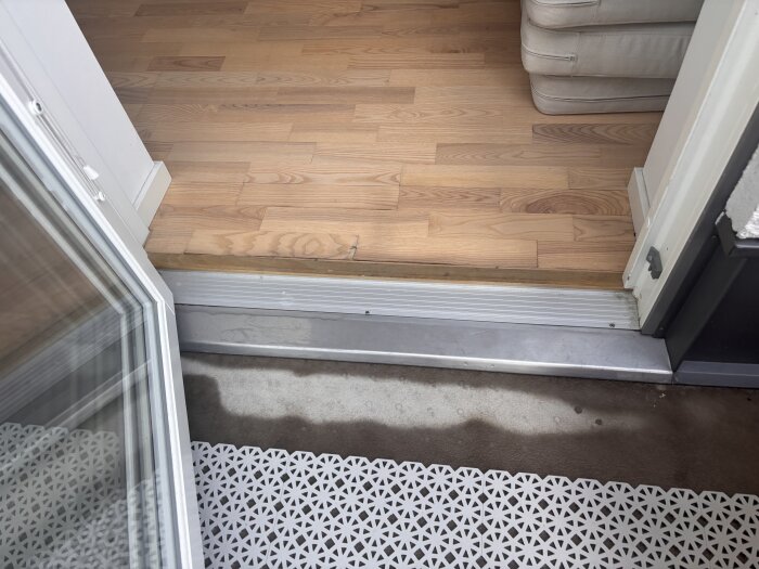 Öppen altandörr med upprest golvbräda inomhus och fuktfläckar på tröskeln.