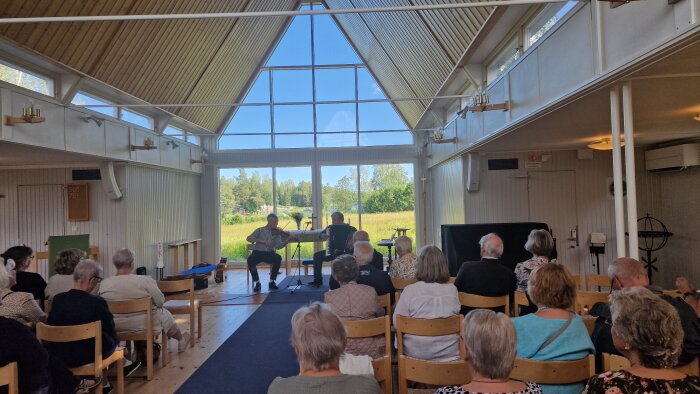En grupp människor sitter i en kyrka och lyssnar på två musiker som spelar fiol och dragspel. Fönstren visar en vy av grönska och blå himmel.