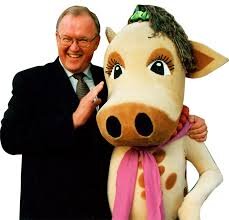 Jag vet inte vem personen är, men bilden visar en man i kostym bredvid en ko-maskot med rosa halsduk och grön hatt.