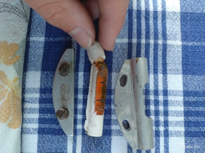 Hand håller ampull med orange vätska från gammal svensk vattenpass, omgiven av två metallbitar på blå-vit-rutig bakgrund.