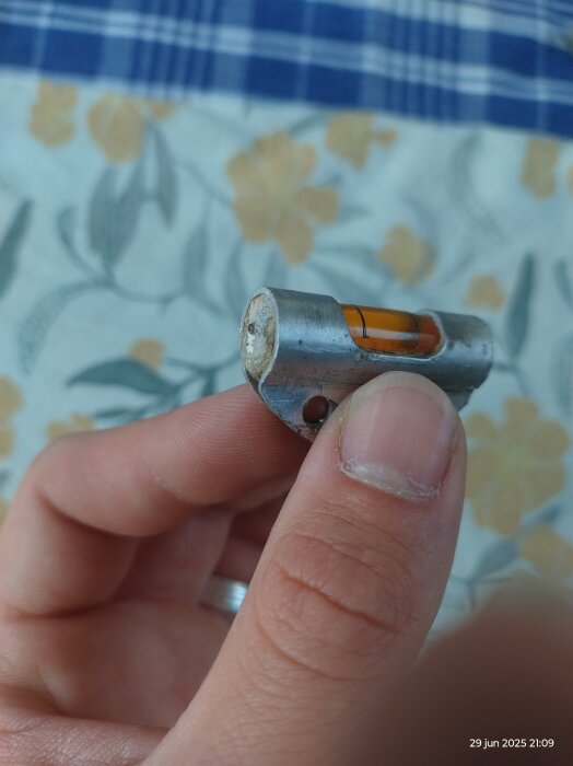 En hand håller i en gammal vattenpassampull med orange vätska i metallhölje, del av en svensk GGB-vattenpass från 1950–60-talet.