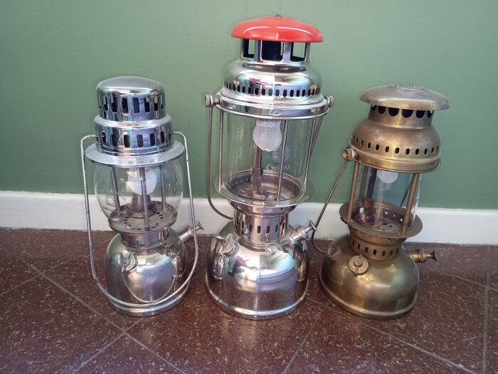 Tre klassiska campinglyktor i metall med olika design, placerade på ett golv mot en grön vägg.