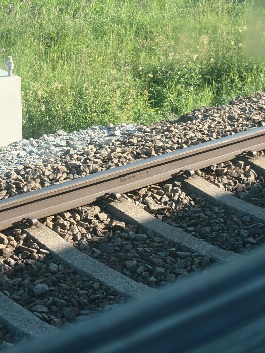 Järnvägsspår med stålbalk, grus och betongslipers, omgiven av grönt gräs i bakgrunden.