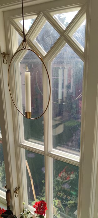 Smutsigt fönster med reflektioner och en hängande ljusstake framför, växter syns utanför.