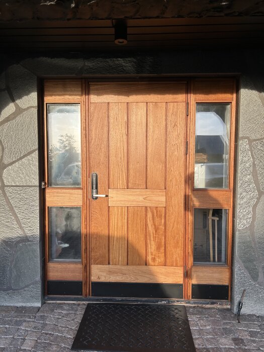 Träytterdörr med glaspaneler, ljusbrun finish och stenram, badar i solens sken.