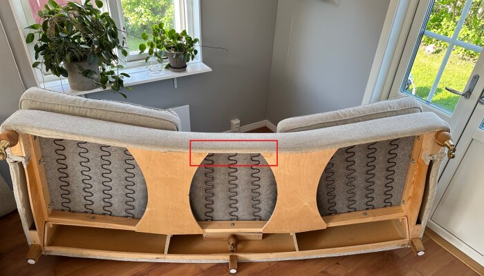 Baksidan av en soffa med avtagna dynor, visar en kloss med spånskiva och fjädrar som behöver omplaceras och limmas om.