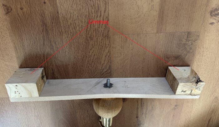 Träplatta med två klossar och en bult, markerade för limning, ligger mot en trägolvbakgrund.