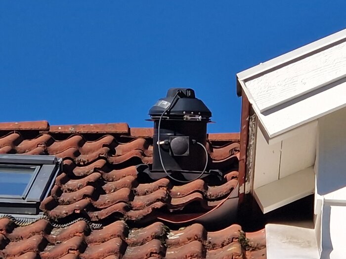 Takfläkt installerad på ett tegeltak intill en takkupa och ett Velux-takfönster, under en klarblå himmel.