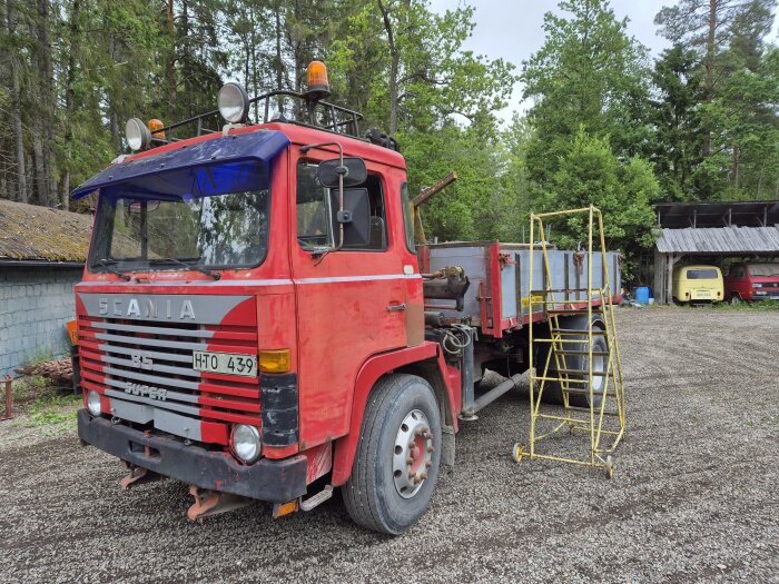 Röd Scania-lastbil med kran och trevägstipp, gul arbetsplattform intill, grusplan och omgivande skog i bakgrunden.
