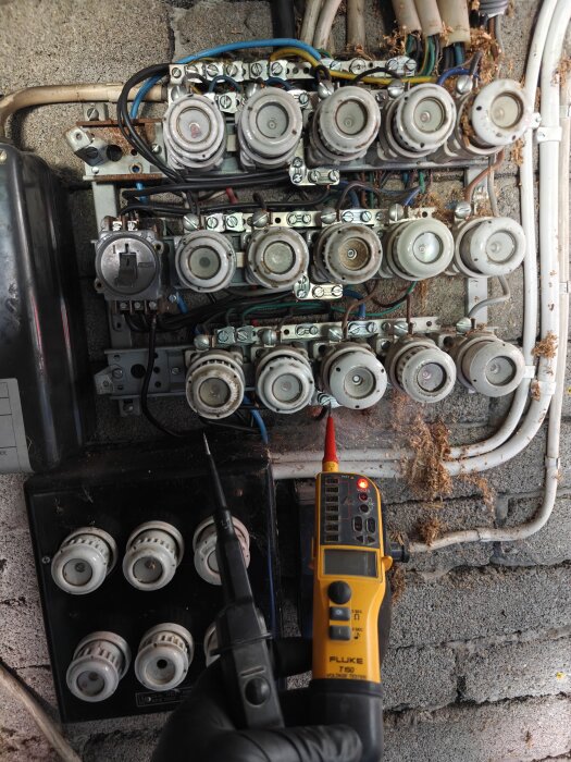 Diazed proppskåp med öppna säkringar och kablar, en hand håller en spänningsmätare nära propparna.