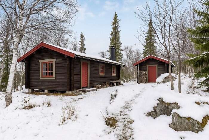 Timmerstuga i snölandskap med rödmålade dörrar och fönster, omgiven av snö och barrträd, inspirerad av traditionell färgsättning.