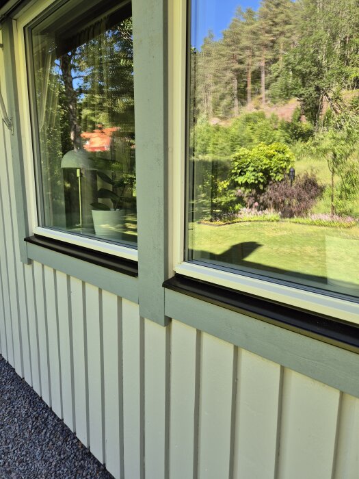 Fönster på ett nyrenoverat hus med ljus träfasad, omgivna av trädgård och grönska i bakgrunden.