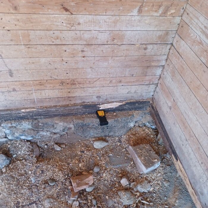 Uthus under renovering med frilagd syll och betonggrund. Träpanel på väggarna, marken täckt med sågspån och stenar. En svart mätare ligger på syllen.