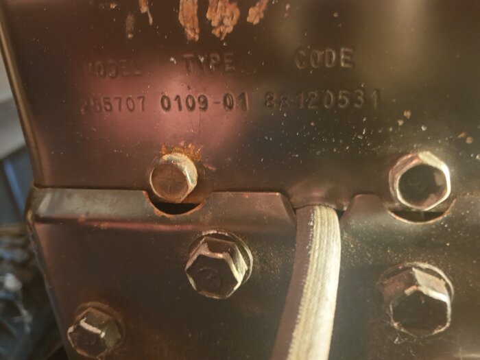 Bild av motornummerplåt med siffrorna 255707, 0109-01 och serienummer 88120531 synliga, omgiven av fyra bultar och en kabel.