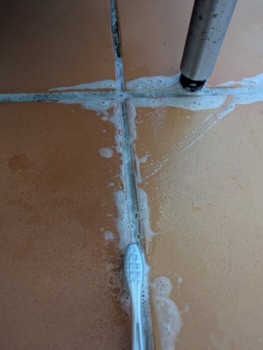 Brun kakelplatta där fogarna rengörs med klorin och tandborste. Visar processen och resultatet av rengöringen.