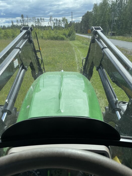 Utsikt från traktorförarhytten över en nyklippt gräsmatta, omgiven av träd och en landsväg i bakgrunden.