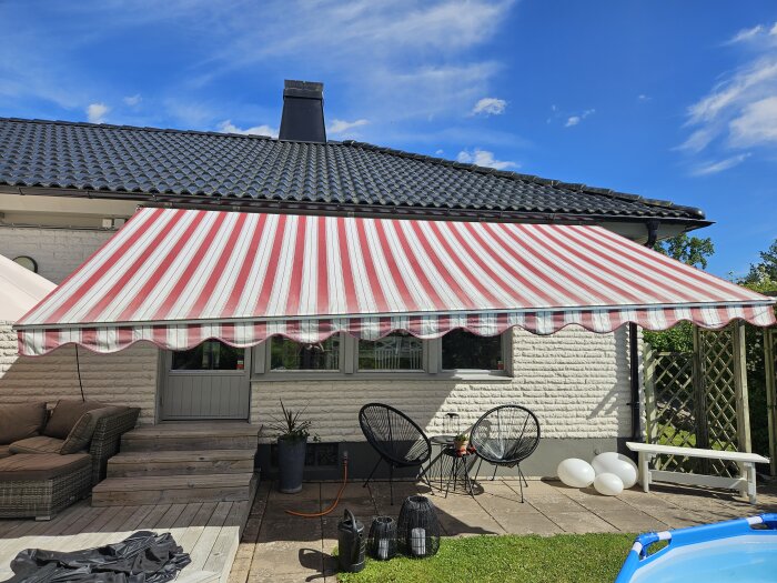 Röd- och vitrandig markis ovanför en uteplats med trappsteg, möbler och en blå barnpool i trädgården.