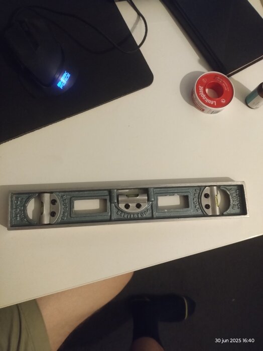 Vattenpass från Eskilstuna med fyrkantiga hål på sidorna, placerat på ett skrivbord nära en datormus och röd tejp.