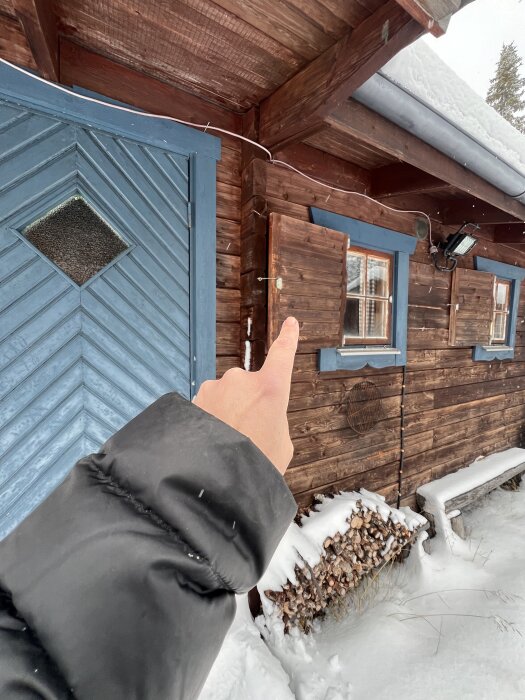 Hand pekar på en brun timmerstuga med blå dörr och blå fönsterkarmar, omgiven av snö och vedstockar.