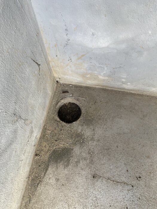 Rund öppning i golv av betong med smuts och sand runt, vid en vitmålad vägg i källartrappa. Möjligen en trasig brunn eller röranslutning.