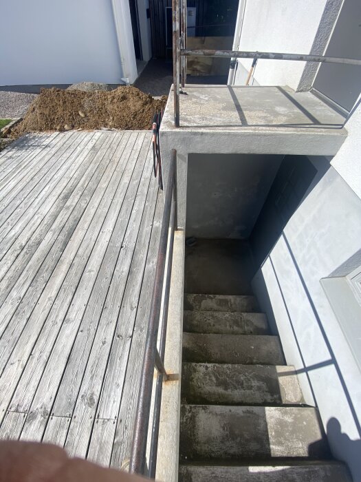 Trappor av betong leder ner till en källardörr, omgivna av ett räcke och en träveranda. En hög med jord ligger närliggande.