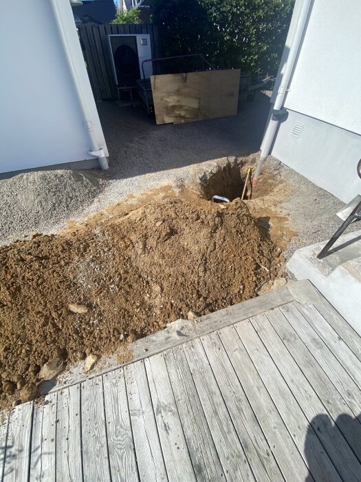 Grävd grop vid husets källartrappa med exponerat rör, omgiven av jord och trätrall, nära en byggnad och staket.