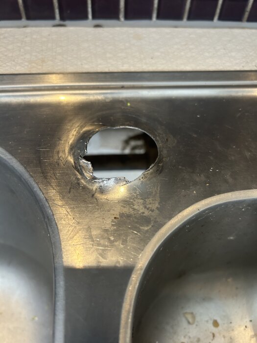 Närbild av diskbänk i metall med ett ojämnt stort hål, efter försök att förstora för en blandare, vilket orsakar glapp mellan blandaren och ytan.