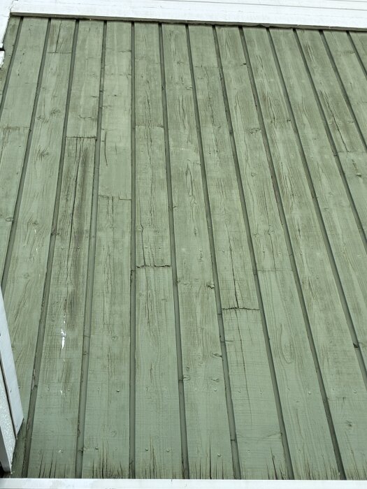 Grön fasad med vertikala brädor, vissa visar tecken på slitage och sprickor.