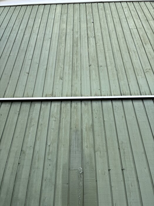 Fasad av gröna lodräta träpaneler på ett hus, vissa brädor är i dåligt skick med synlig slitage och ålderstecken.