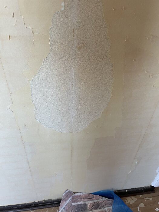 Murstock med rester av tegeltapet kvar efter borttagning, synbara lager och avskavda delar. Kasserad tapetremsa på golvet.