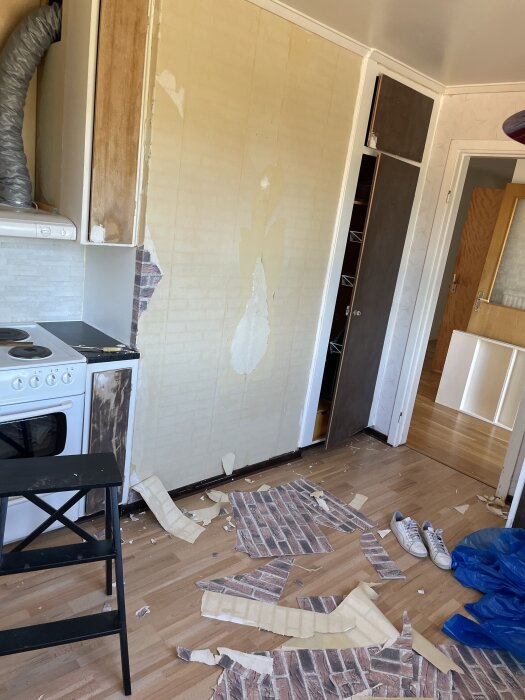 Vägg med borttagen tegeltapet, kvarvarande lager syns. Tegeltapetbitar ligger på golvet, samt en pall och ett par skor.