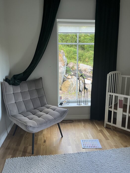 Rum med stort fönster utan foder, fåtölj, mörka gardiner och vit barnsäng. Utsikt mot trädgård med stenar.