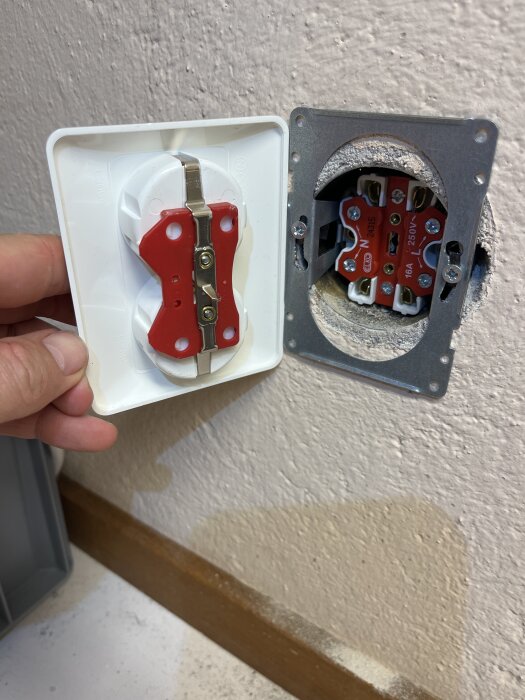 Öppen vägguttaginstallation med röd baksida och metallram där framsidan av uttaget hålls vid sidan av en hand.