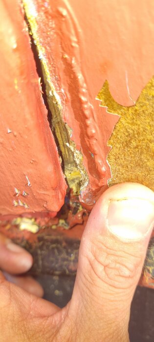 Närbild på en skadad och flagnande yta med färg i rött, där det underliggande materialet är exponerat. En hand håller i ytan.