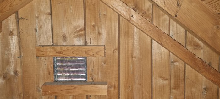 Träpanel på vinden med masonit fastsatt, synlig ventilation öppning i panelen.