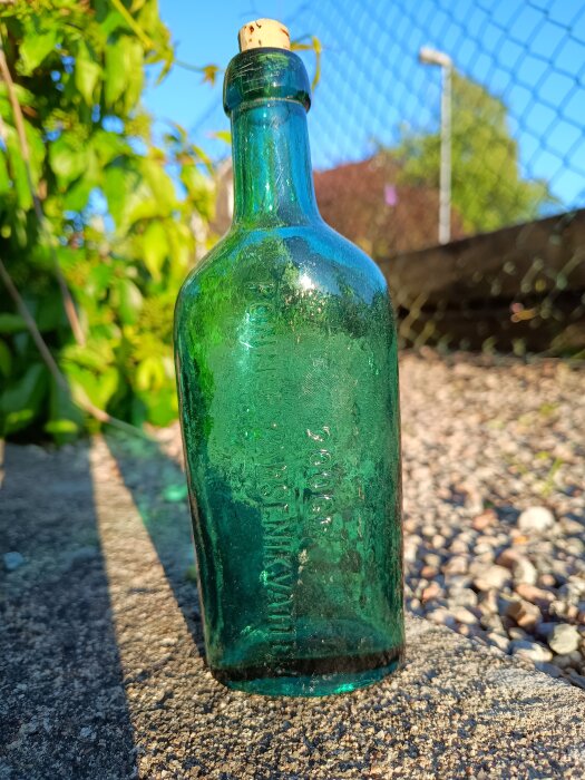 Grön glasflaska med texten "Ronneby arsenikvatten", stående utomhus mot en bakgrund av stängsel och grönska.