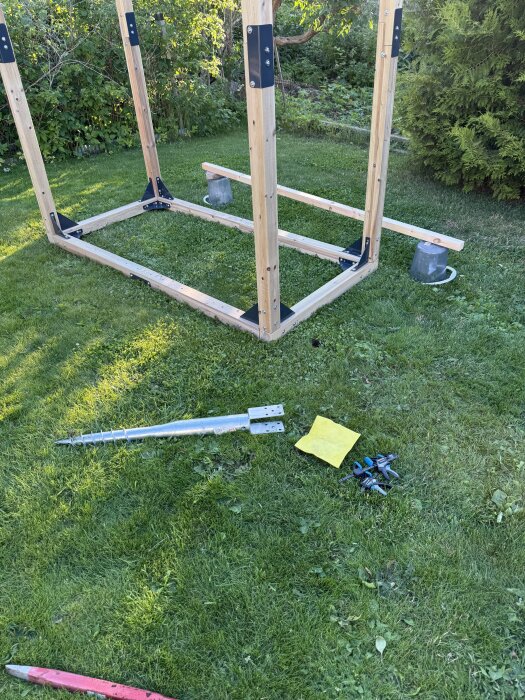 Träram för klätterställning ligger på gräsmatta; markskruv, spett, gula trasor och andra verktyg ligger bredvid.