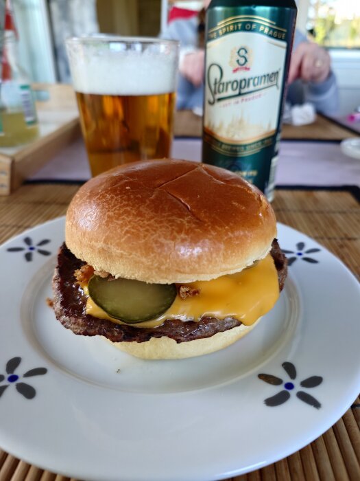 Hamburgare med ost och gurkskivor pÄ en tallrik, bredvid ett glas öl och en burk Staropramen-öl.