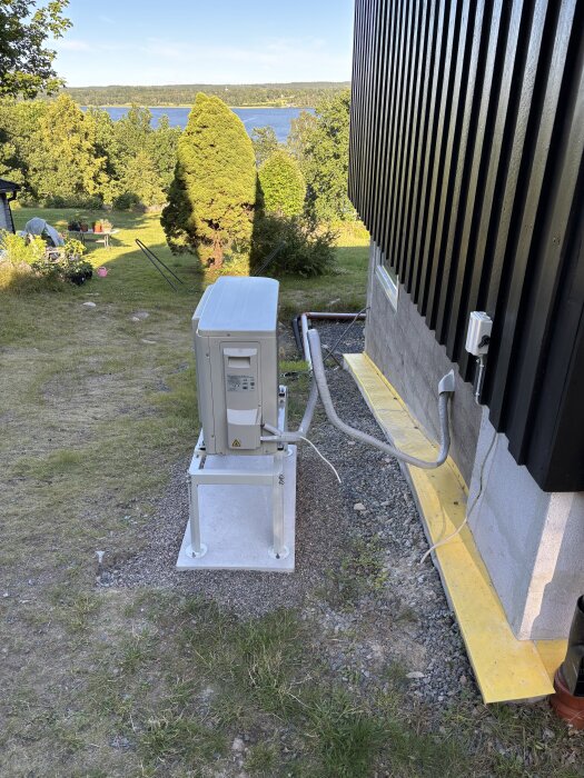 Luft-luft värmepump på stativ nära en husvägg med sladd i en loop bakom, placerad på en gjuten plint med trädgård i bakgrunden.
