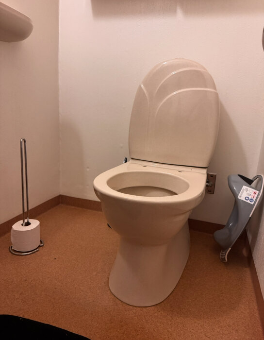 Toalettstol märkt "Gustavsberg 82" och "380" i ett badrum, med ett papperställ bredvid. Ena sitsarmen saknas.