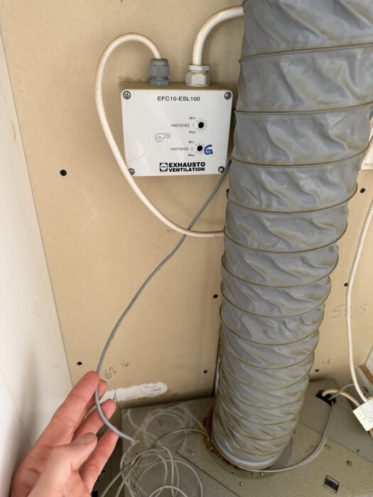 Styrdonet EFC10-ESL100 från Exhausto Ventilation med anslutna kablar och en luftslang syns på bilden. En hand håller i en lös kabel.