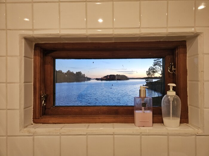 Utsikt över en sjö och öar genom ett fönster med två tvålpumpar på fönsterkarmen, kaklade väggar runt omkring, himlen i solnedgång.