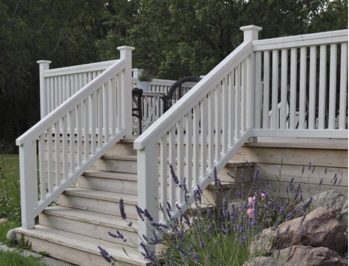 Vit målad trappa med räcke och spjälor, omgiven av stenar och lavendel i en trädgårdsmiljö.