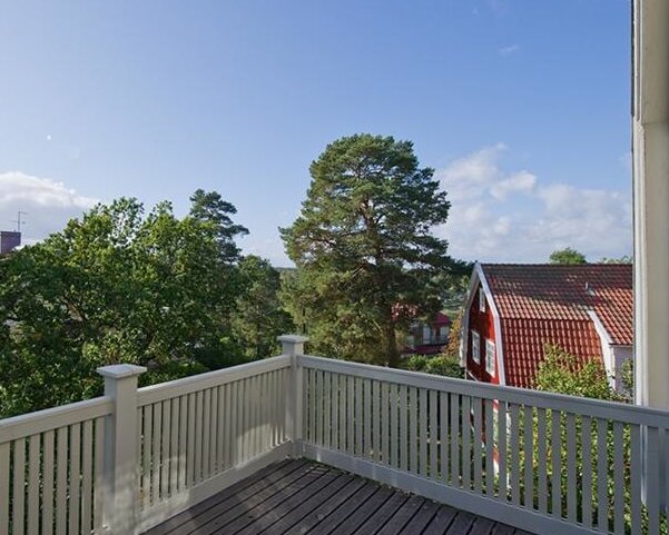 Vitt altanräcke med smala spjälor och träpelare, omgiven av gröna träd och ett rött hus med brant tak i bakgrunden.